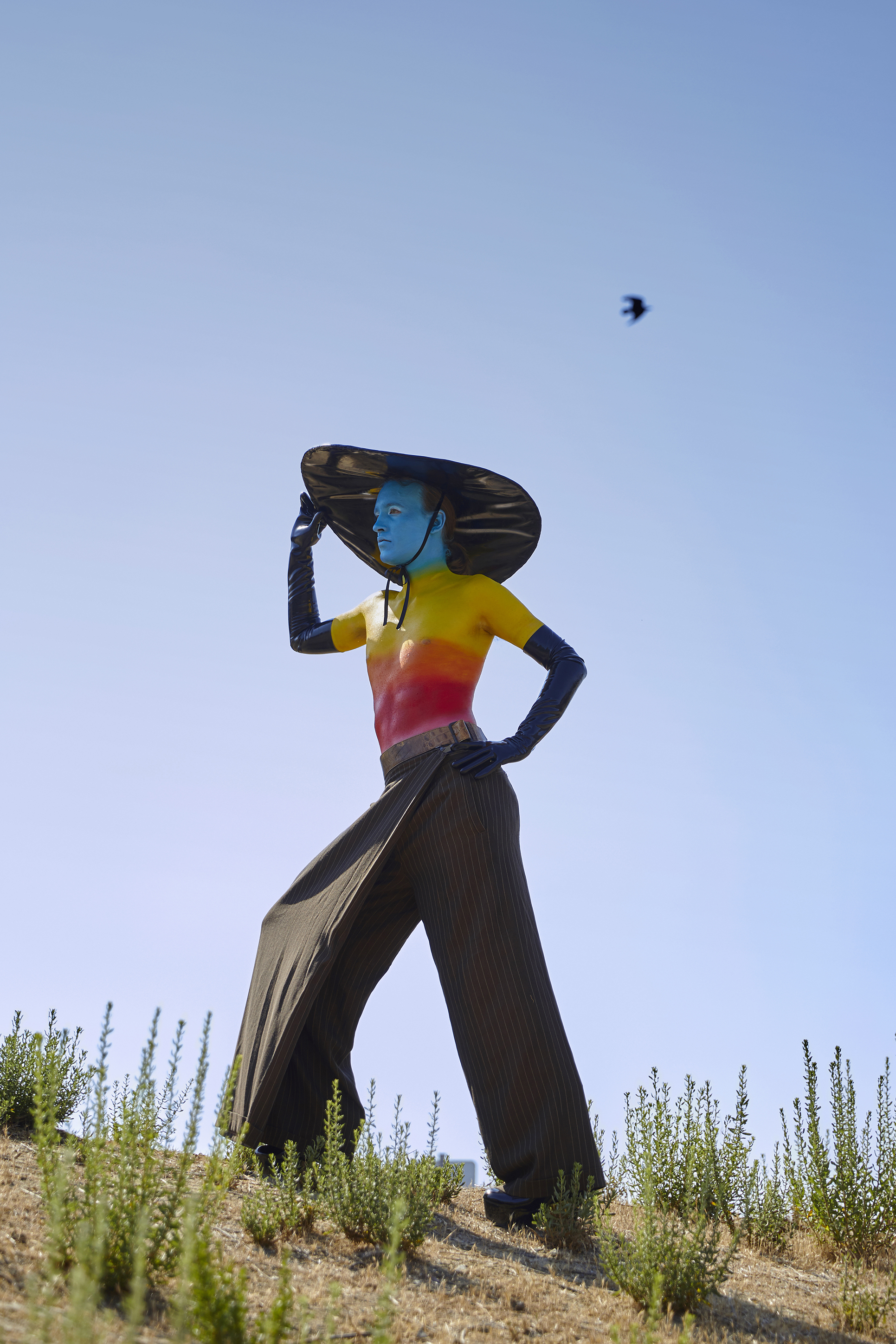 Jesse shirtless with rainbow body paint, latex hat and gloves on a hill outside posing